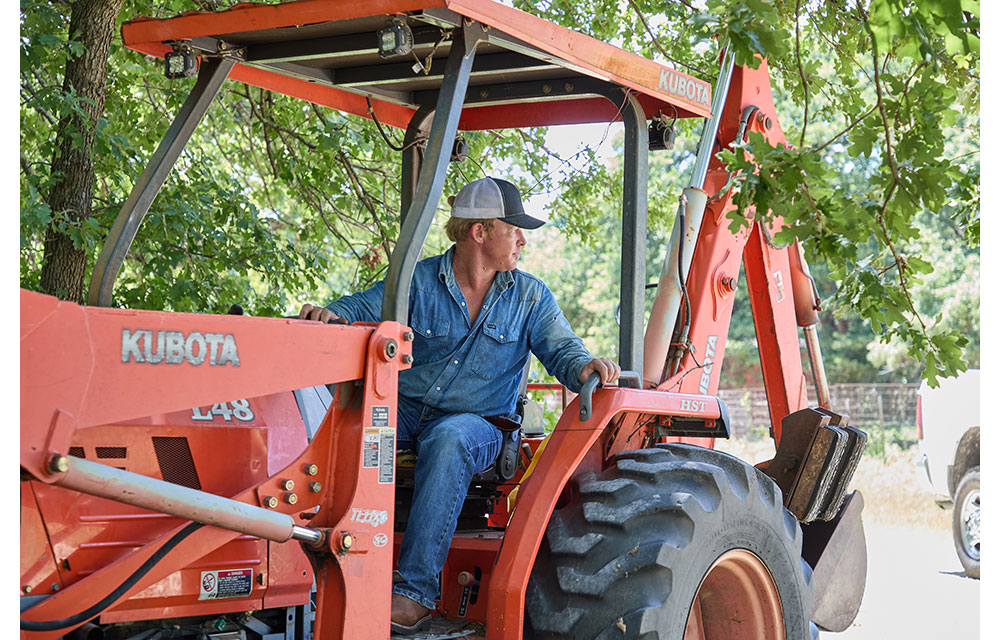 Un hombre, Wes Stevenson, con una camisa de mezclilla y una gorra, está sentado en un tractor naranja bajo árboles frondosos, mirando hacia un lado mientras opera el equipo.